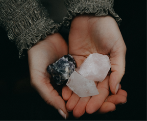 Hand met drie ruwe edelstenen (amethist, rozenkwarts, bergkristal) op een donkere achtergrond